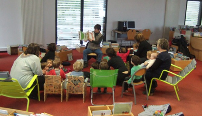 Accueil de petite enfance à la bibliothèque d'Acigné. Le bibliothécaire montre le livre qu'il conte à un groupe d'enfants en rond sur des tapis