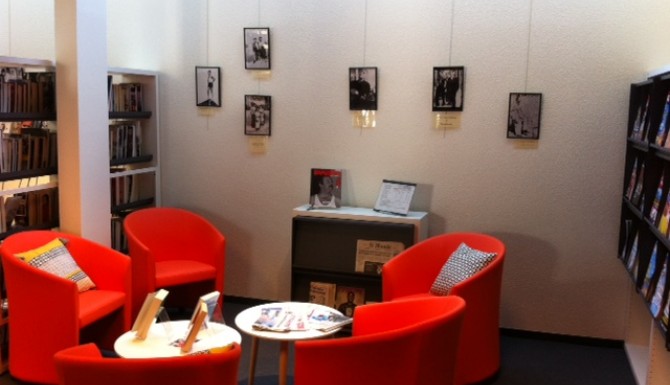 Salon de lecture avec des fauteuils cosy rouges et tables basses. Au fonds, on voit une exposition