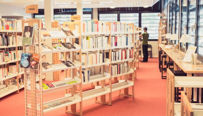 autre vue des mêmes espaces. On y distingue des tables de lecture hautes le long des vitres, munies de lampes de chevet individuelles