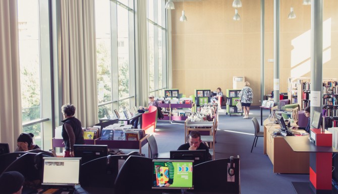 au premier plan l'espace multimedia, des ordinateurs en vis à vis. En arrière-plan, l'automate de prêt de couleur rouge, et des bacs d'albums et BD de couleur violette et verte.