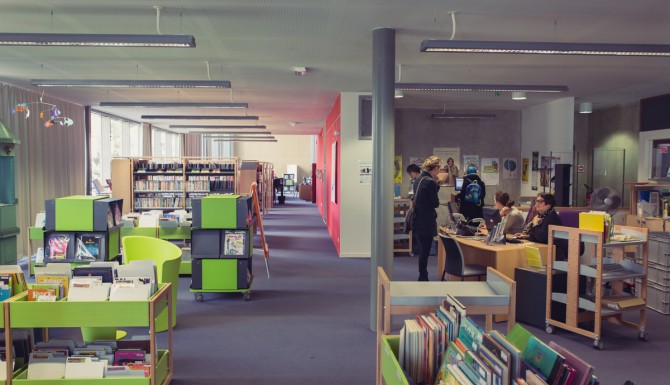 espaces de la bibliothèque. On y aperçois l'accueil, des bacs de couleur verte contenant des BD et des albums. Murs rouges et moquette mauve