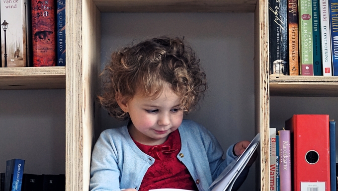 Reading (CC BY-NC 2.0) by Erik Schepers Une petite fille lit un livre assise dans le bas d'une bibliothèque en bois.