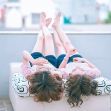 Deux femmes côte à côte sont allongées sur un lit et nous tournent le dos. Leurs pieds sont posés sur un muret
